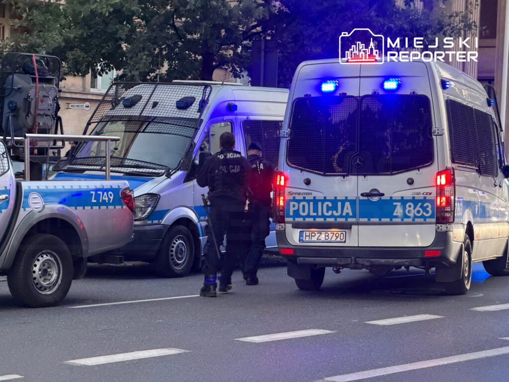 Grupa policjantów w umundurowaniu, z radiowozami na tle ulicy, prowadzi działania interwencyjne w mieście.