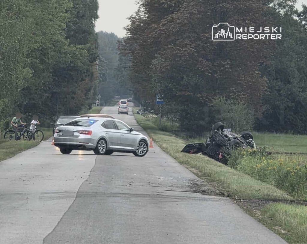 Na drodze stoi srebrny samochód policyjny z włączonymi sygnałami, a obok dwóch rowerzystów obserwuje przewrócony motocykl w rowie.
