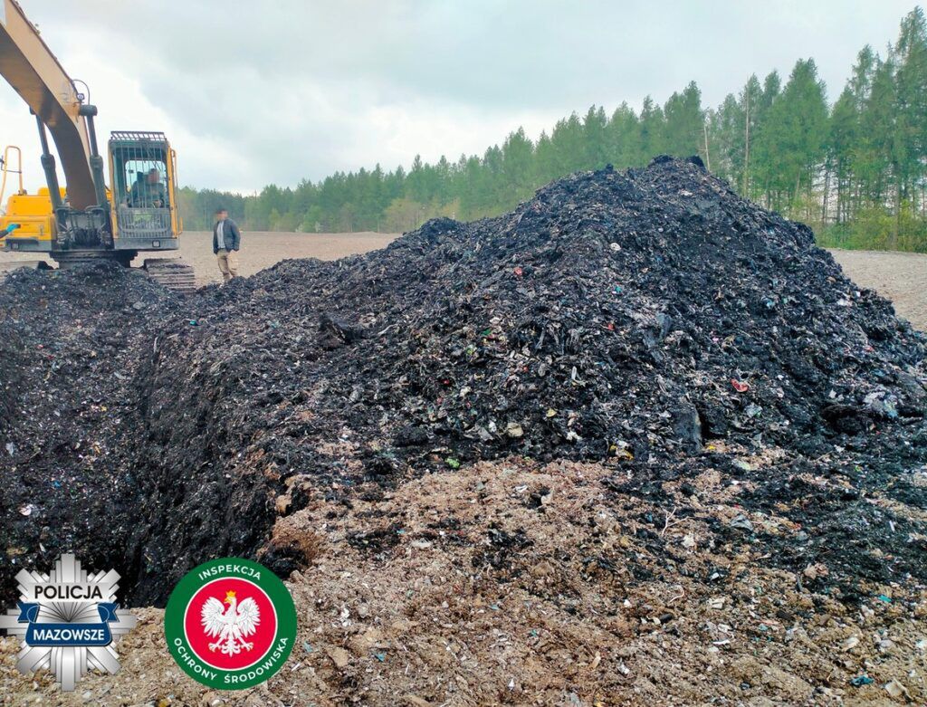 Mężczyzna w roboczym ubraniu stoi obok koparki, przy górze czarnego odpadu w lesie.
