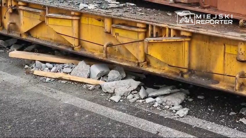 Uszkodzony fragment żółtego wagonu kolejowego z odłamkami betonu i drewnianymi belkami leżącymi na drodze.