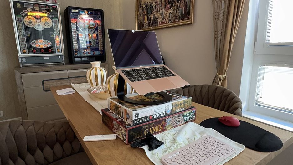 Laptop umieszczony na stosie gier planszowych, obok myszka komputerowa i różowa klawiatura, w jasnym wnętrzu z automatami do gier.