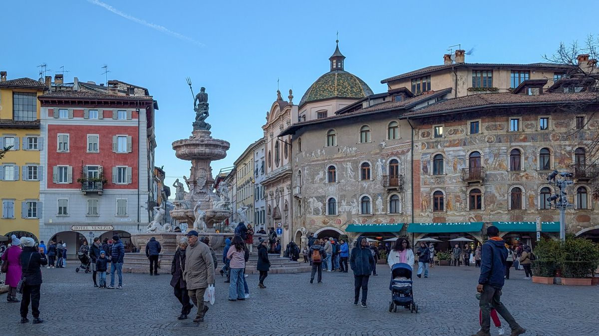 A crowd of people gathers in the cobblestone Piazza Duomo, surrounding the historic Fountain of Neptune (Fontana del Nettuno), with the Duomo bell tower and colorful buildings forming the backdrop in Trento, Trentino, Italy, on November 23, 2025. The city square is a central point for daily life and is also a key location along the route for the Olympic Torch Relay, scheduled for January 29, 2026, for the Milano Cortina 2026 Winter Olympics. (Photo by Michael Nguyen/NurPhoto via Getty Images)