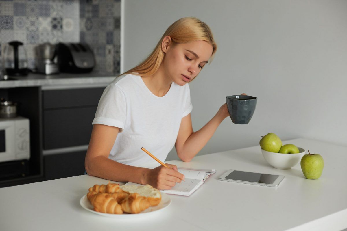 Young blonde girl planning her day, keeps a big grey cup, writes plans in a dairy. Croissants on a plate, apples, are there on a white kitchen table, kitchen background