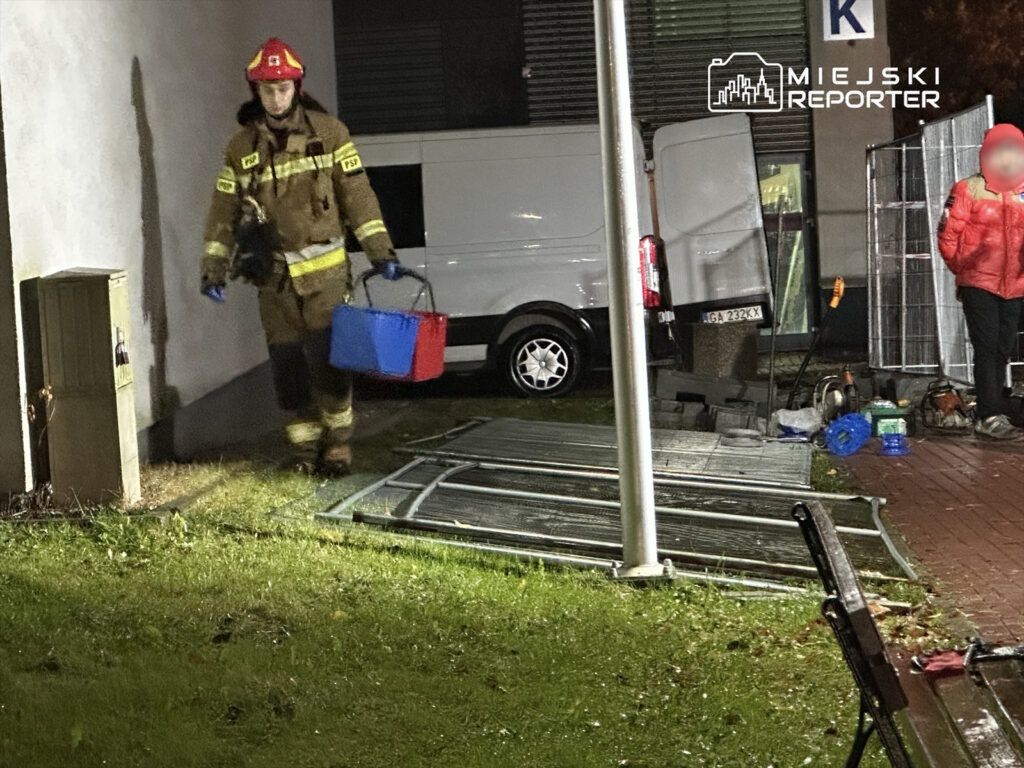 Strażak w mundurze niesie niebieską i czerwoną torbę w pobliżu budynku, obok zaparkowanego samochodu dostawczego.
