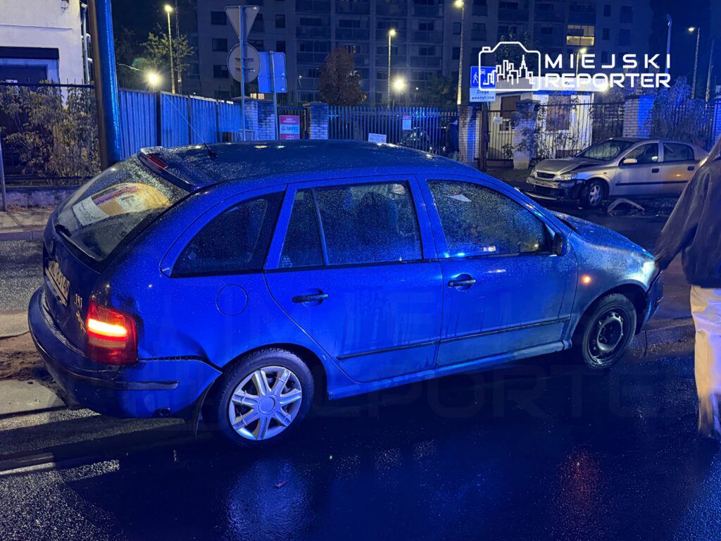 Niebieski samochód osobowy zaparkowany na poboczu drogi, z uszkodzeniem tyłu, w nocy przy oświetlonych budynkach.