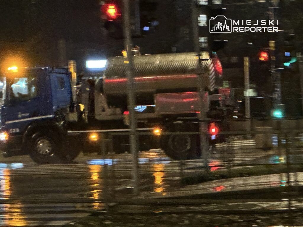 Niebieski samochód ciężarowy z cysterną przejeżdża przez mokrą ulicę w deszczową noc, mijając sygnalizację świetlną.