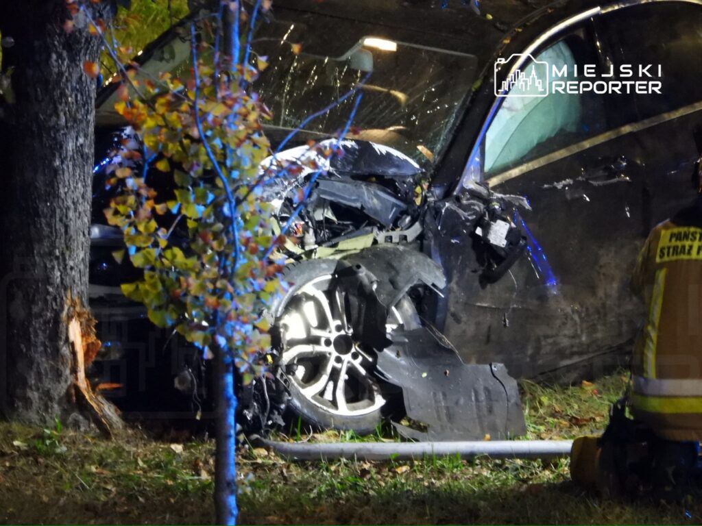 Wgnieciony samochód osobowy uderzony w drzewo, z widocznymi uszkodzeniami nadwozia, w otoczeniu służb ratunkowych.
