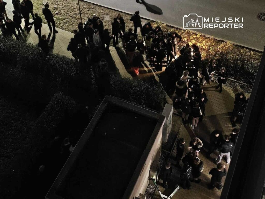 Grupa młodych ludzi stoi na chodniku, rozmawiając i spędzając czas w nocnej scenerii. W tle widoczna jest oświetlona roślinność.