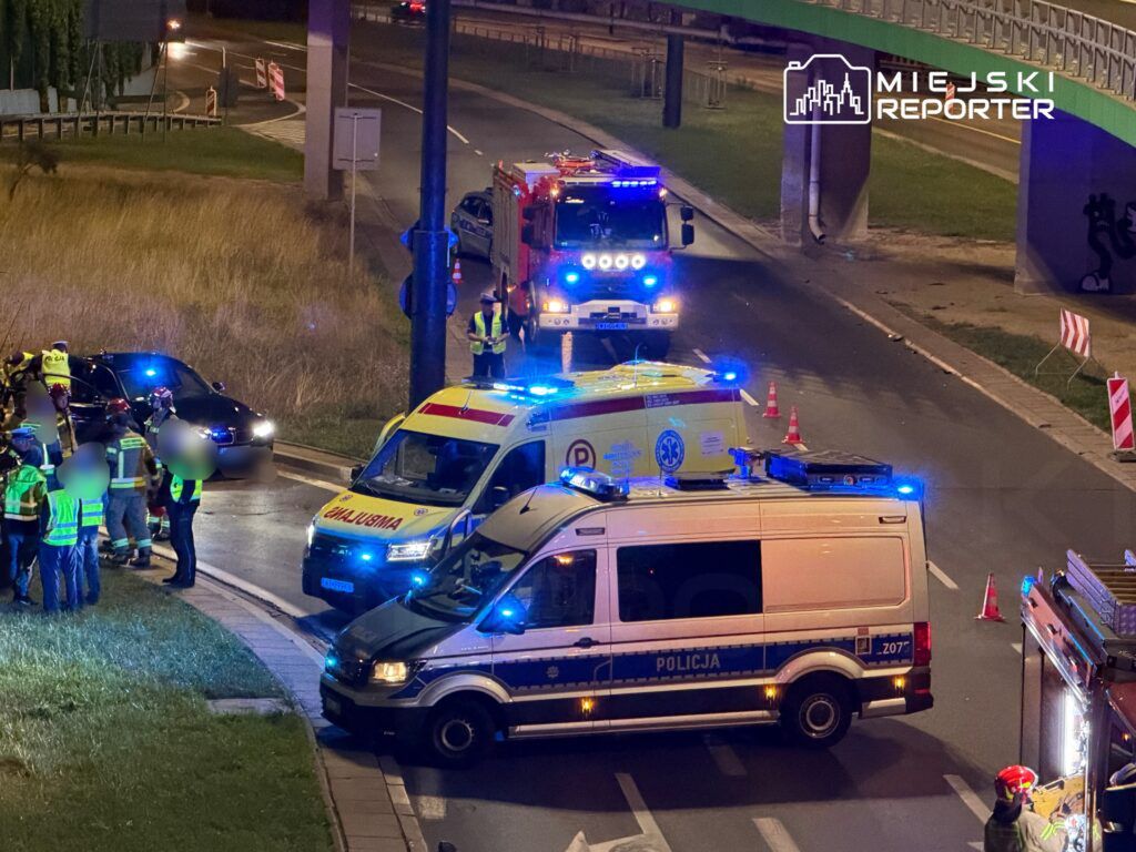 Grupa ratowników i policjantów prowadzi działania na miejscu wypadku, obok zaparkowanych pojazdów służb ratunkowych.