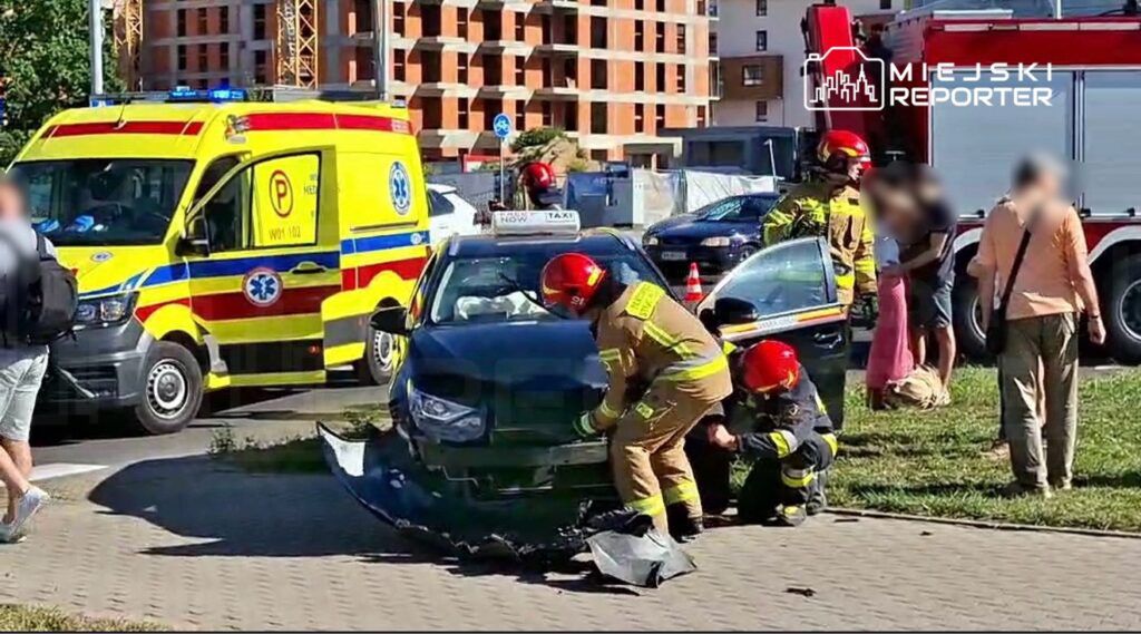 Ratownicy medyczni udzielają pomocy poszkodowanemu w wypadku drogowym przy nowym budynku, obok karetki i straży pożarnej.