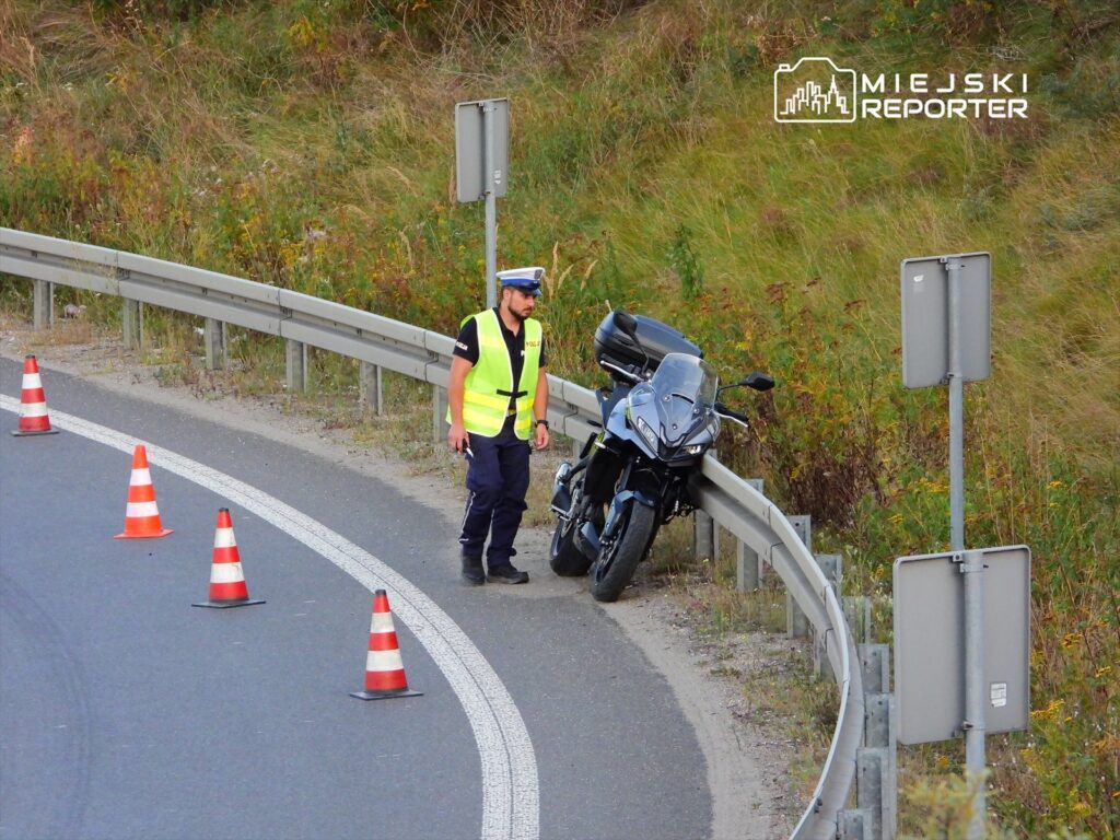 Policjant w odblaskowej kamizelce kontroluje motocykl zaparkowany przy drodze, otoczony pachołkami.