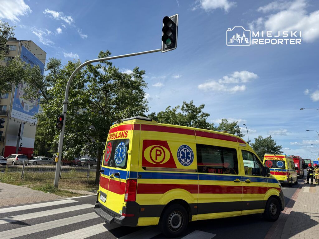Karetka pogotowia zatrzymana na czerwonym świetle na pasach, obok innych pojazdów w miejskim otoczeniu.