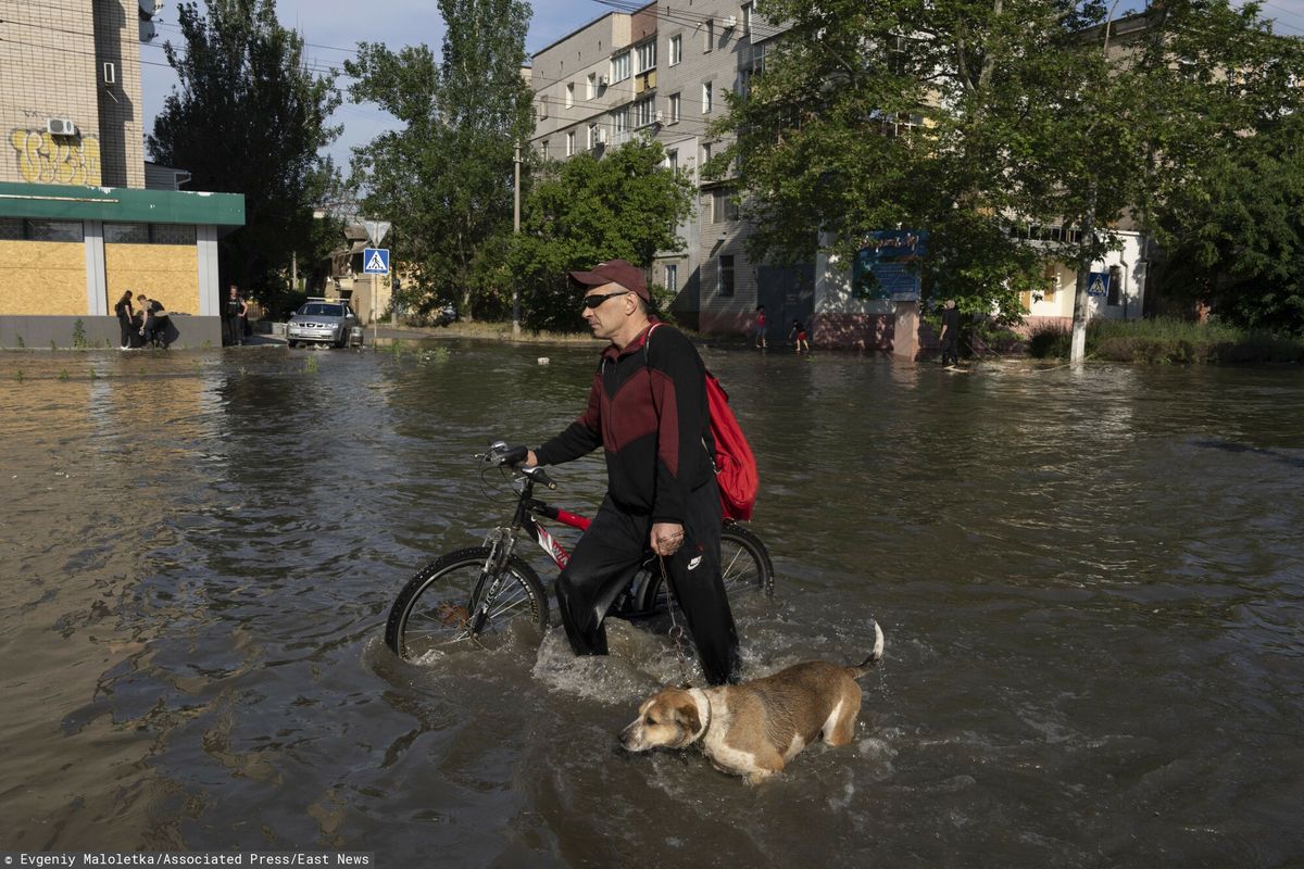 Lokalny mieszkaniec z rowerem i psem idzie ulicą obok budynków w Chersoniu na Ukrainie, wtorek, 6 czerwca 2023 r., które zostały zalane po wysadzeniu zapory w Nowej Kachowce w nocy.