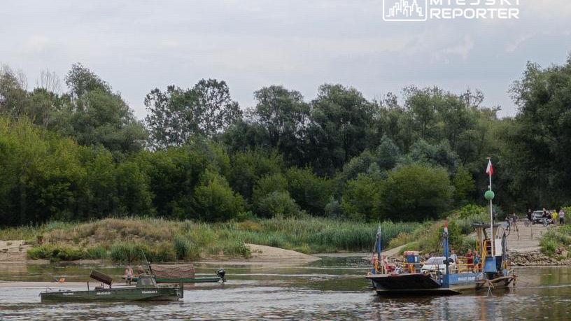 Dwie łodzie na rzece, jedna z pasażerami, druga z silnikiem, otoczone zielonymi drzewami i piaszczystym brzegiem.