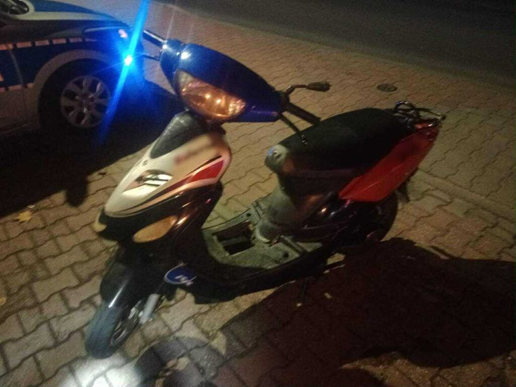 Skuter zaparkowany na kostkowanej nawierzchni obok radiowozu policyjnego z włączonymi światłami.