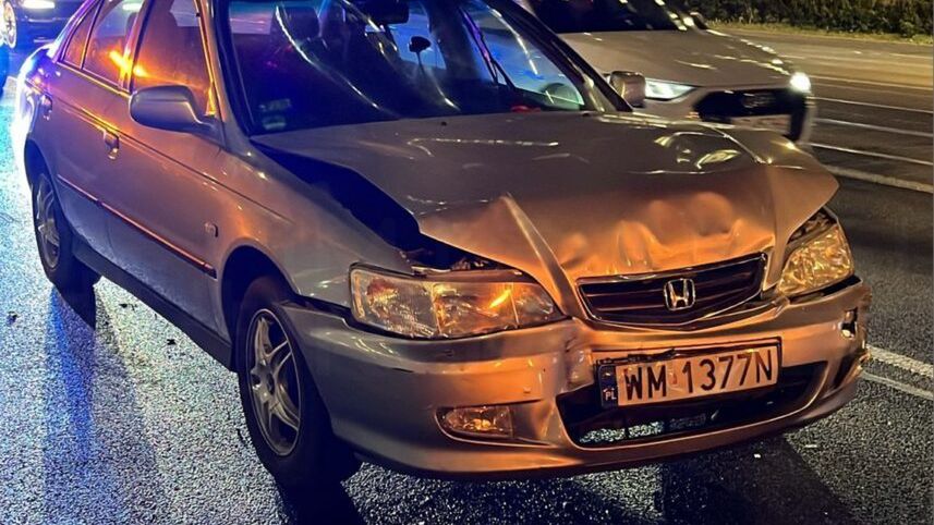 Uszkodzony samochód osobowy marki Honda z wgniecionym przodem stoi na mokrej jezdni w miejskim otoczeniu.