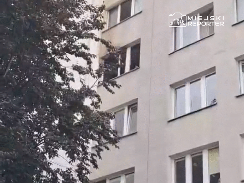 Mężczyzna obserwujący otoczenie z okna mieszkania w bloku, w tle drzewa i elewacja budynku.