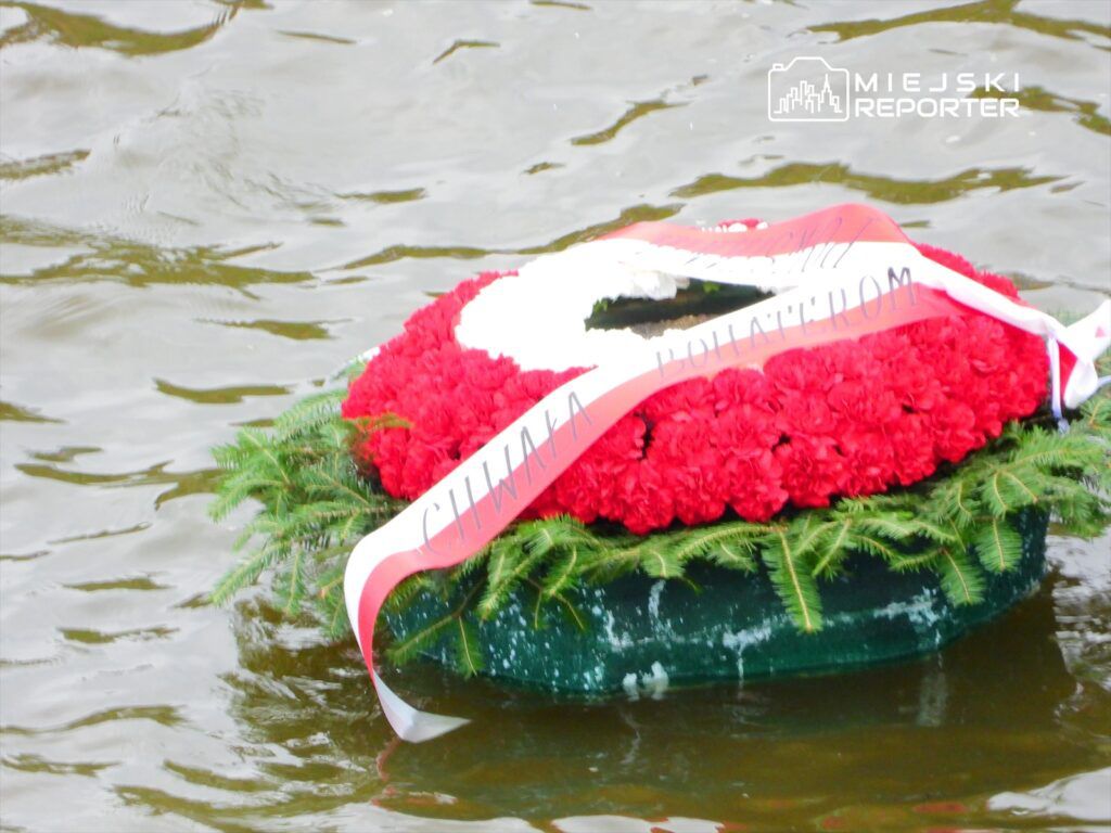 Wieniec z czerwonymi kwiatami i wstążką unoszący się na wodzie.