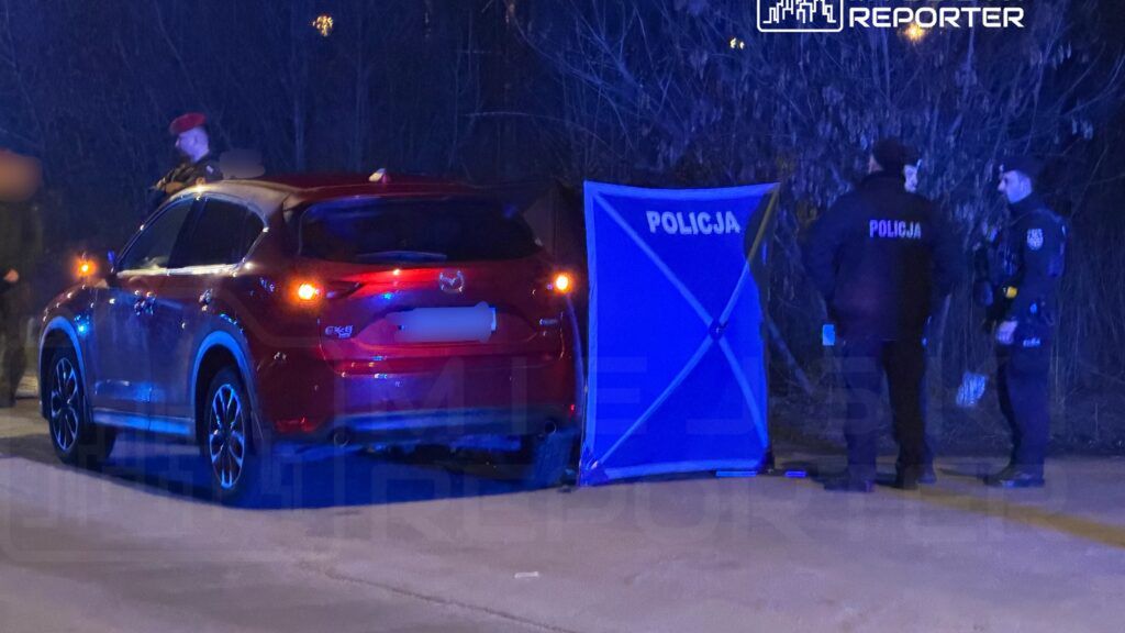 Czerwony samochód osobowy zatrzymany przy drodze, obok policjanci rozmawiający przy niebieskiej barierce z napisem "POLICJA".