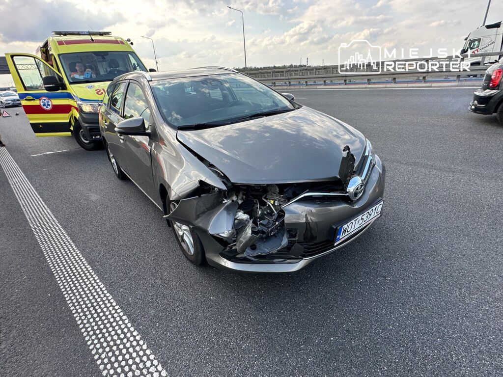 Uszkodzony samochód osobowy w kolorze szarym stojący na drodze, obok karetka pogotowia.