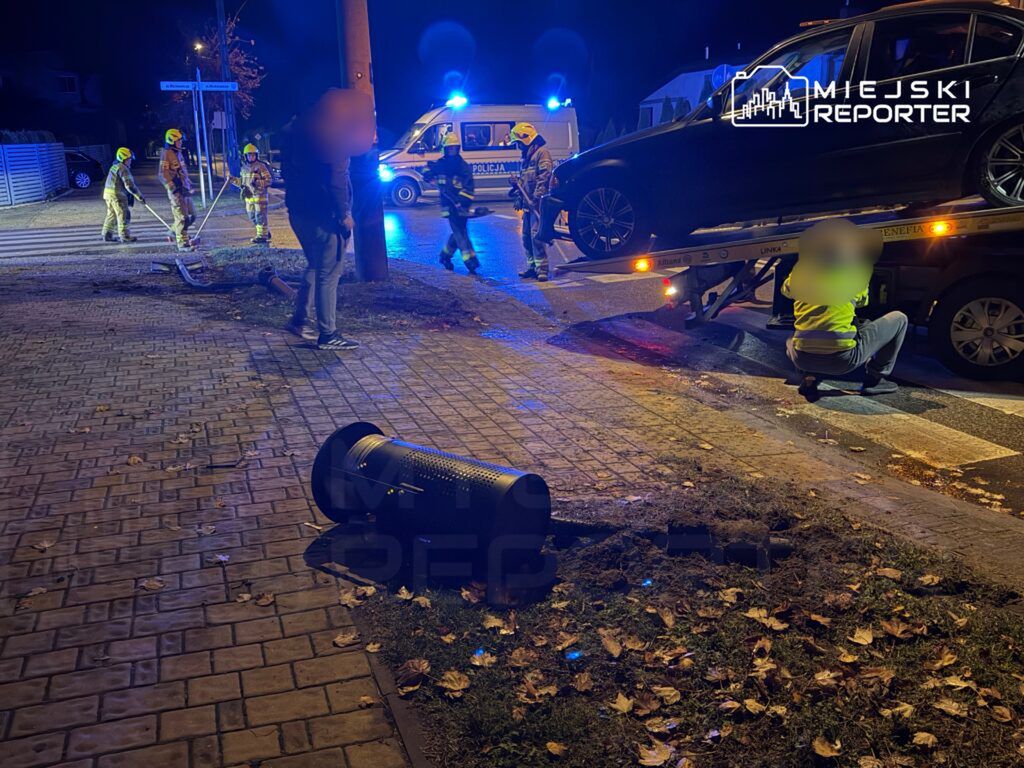 Grupa strażaków pracuje przy przewróconym koszu na śmieci, obok stoi policyjny radiowóz, a auto jest wciągane na lawetę.