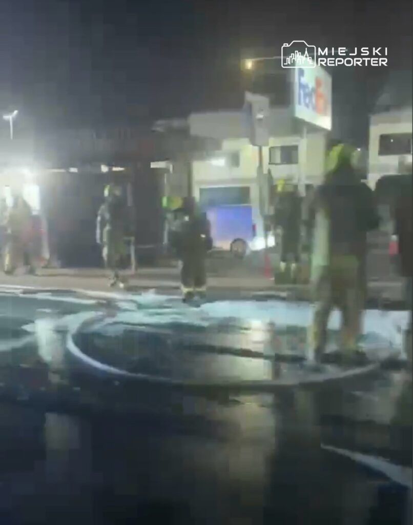 Grupa strażaków w hełmach pracuje nocą na miejscu akcji, oblewając podłoże pianą gaśniczą.