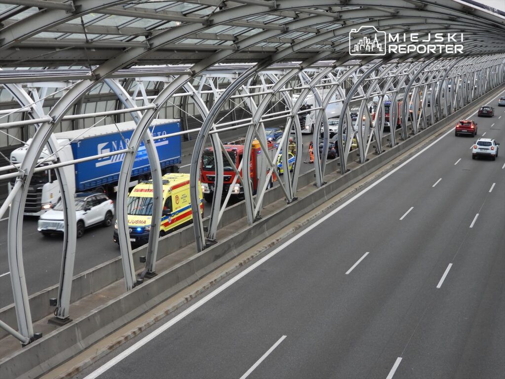 Karetka i pojazdy służb ratunkowych zatrzymane na pasie drogowym, obok ruchu samochodowego na autostradzie.