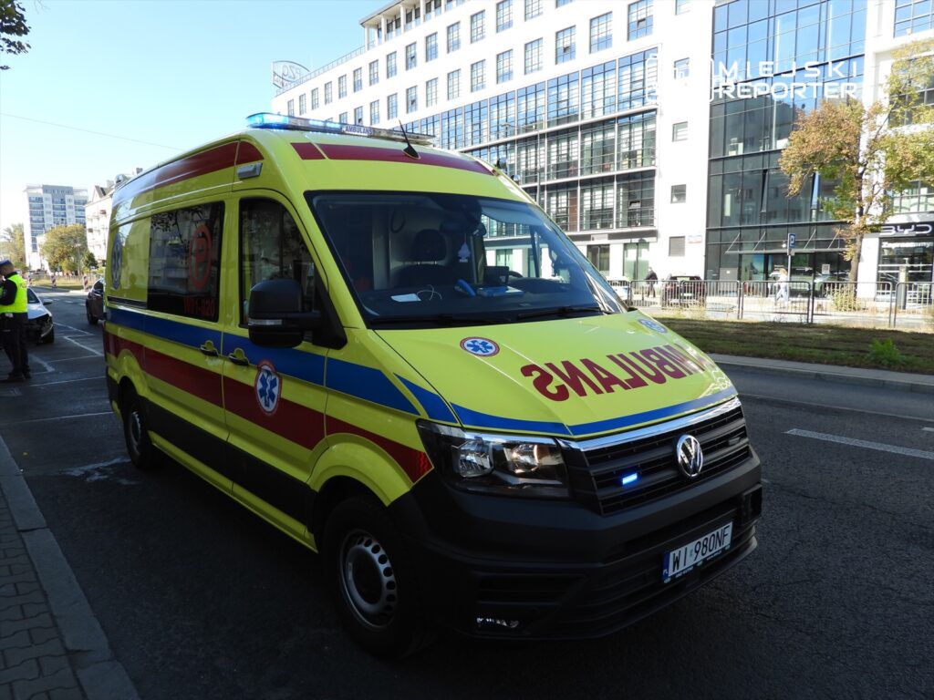 Ambulans w żółto-czerwonych barwach zatrzymany na ulicy w mieście, z włączonymi sygnałami świetlnymi.