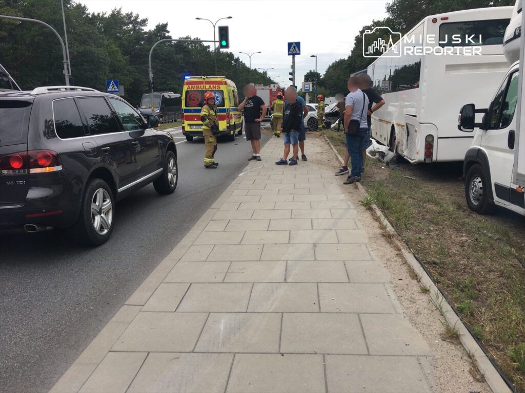 Grupa osób stojąca obok drogi, w tle widać karetki pogotowia oraz strażaka rozmawiającego z mężczyzną.