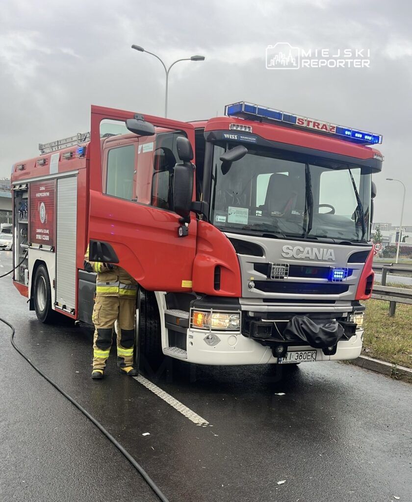 Strażak w mundurze obsługuje wóz strażacki marki Scania na mokrej nawierzchni drogi.