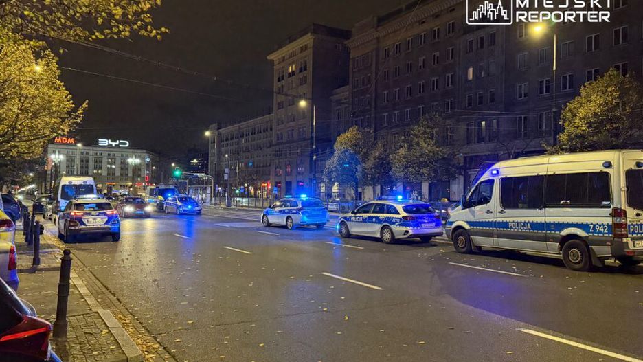 Policyjne samochody z włączonymi sygnałami świetlnymi zatrzymane na ruchliwej ulicy nocą, w tle oświetlony budynek.