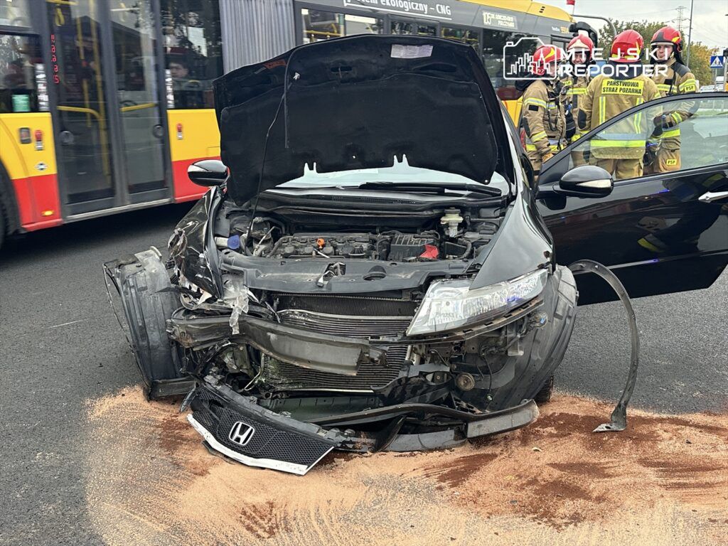 Czarny samochód osobowy z uszkodzoną przednią częścią stoi na drodze, obok grupy strażaków.
