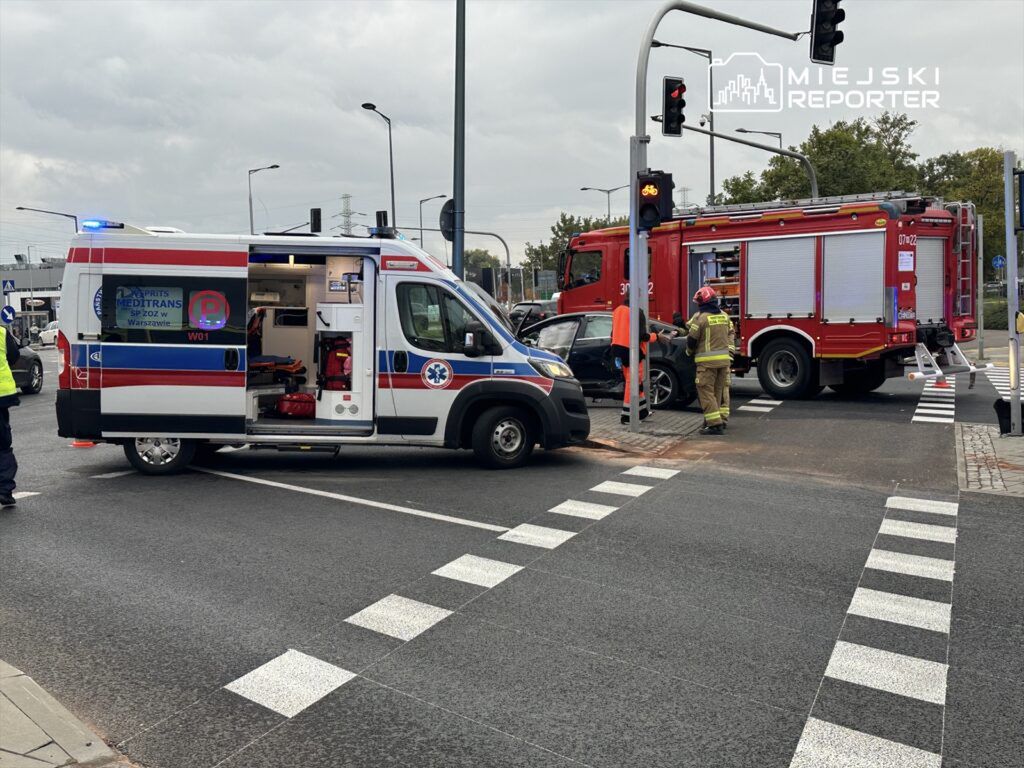 Karetka pogotowia i wóz strażacki interweniują na skrzyżowaniu po wypadku drogowym. Ratownicy pomagają poszkodowanemu.