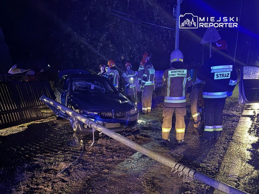 Grupa strażaków w mundurach ocenia uszkodzone auto przy przewróconym słupie energetycznym w deszczowym otoczeniu.