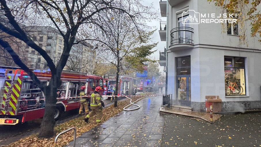 Strażacy gaszą pożar przy budynku mieszkalnym, obok stoją wozy strażackie i wąż gaśniczy na chodniku.