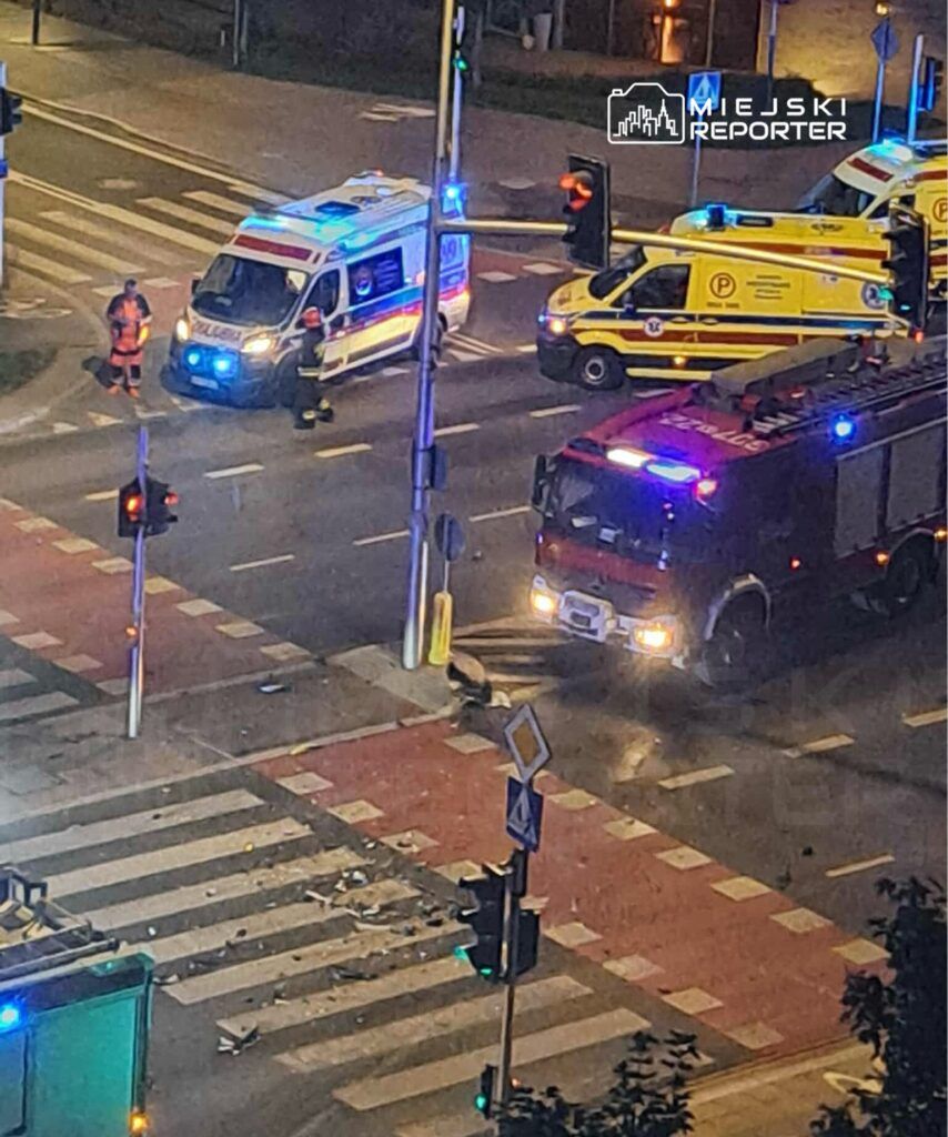 Na skrzyżowaniu ulicy widać pojazdy ratunkowe z włączonymi sygnałami świetlnymi oraz straż pożarną. W tle leżą fragmenty rozbitego pojazdu.