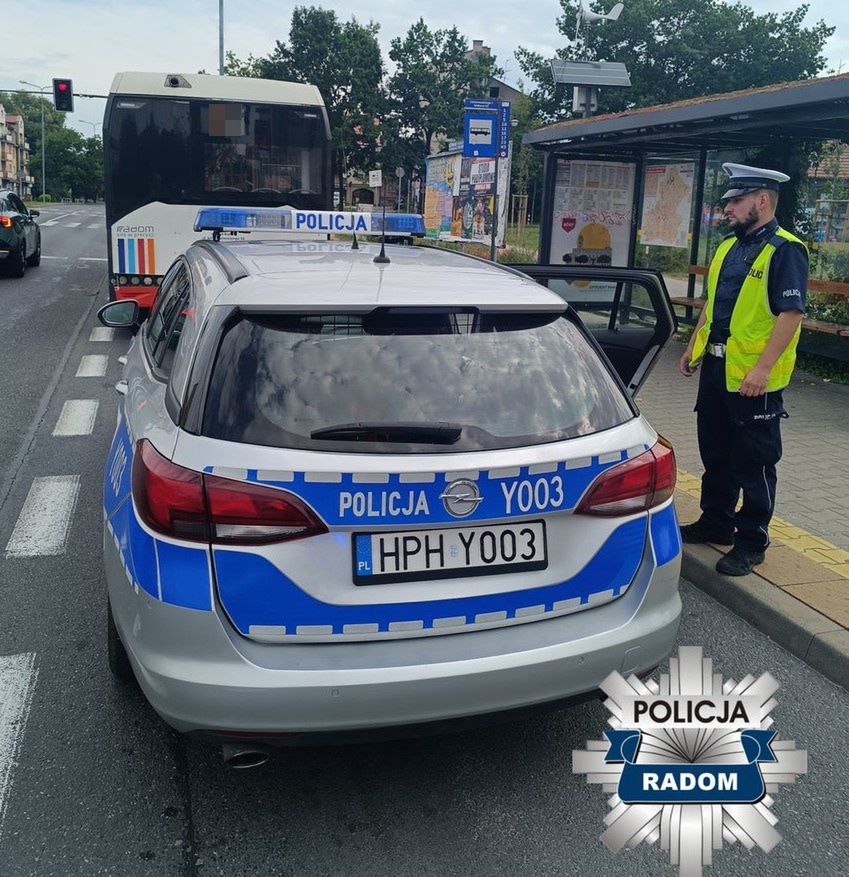 Policyjny radiowóz zatrzymany przy przystanku autobusowym, obok funkcjonariusz rozmawia z kierowcą.