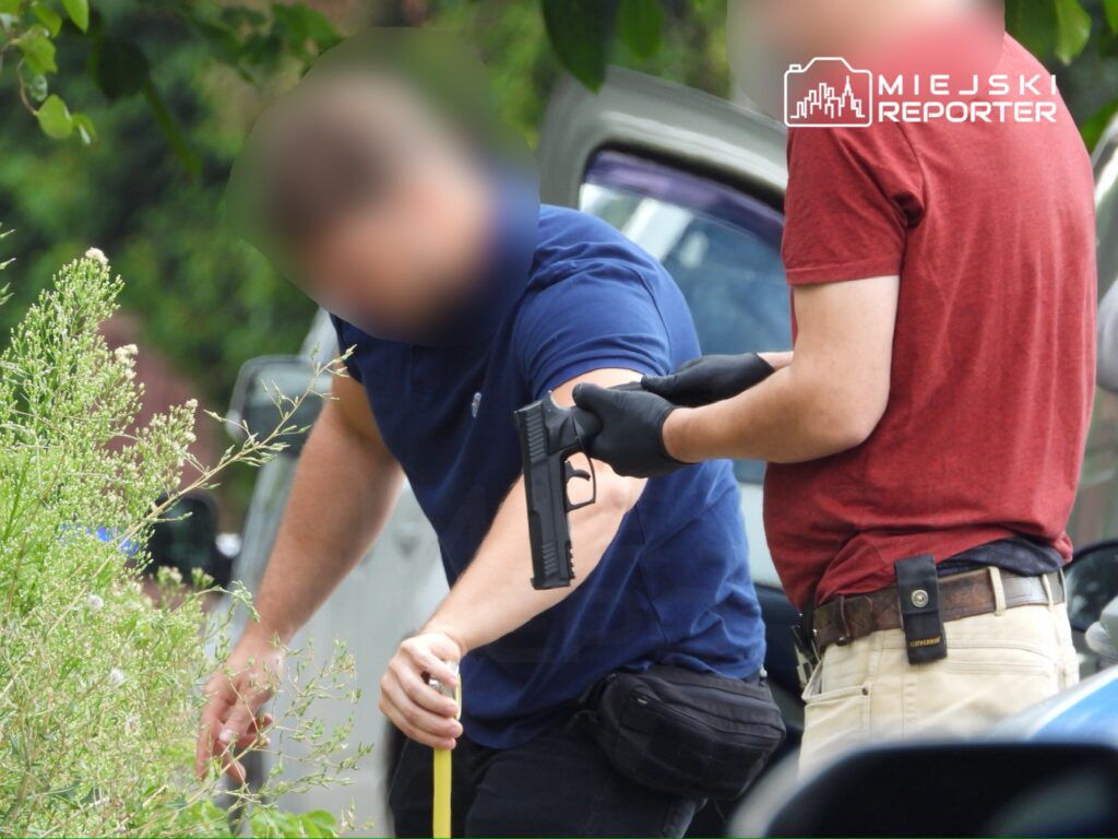 Mężczyzna w granatowej koszulce trzyma miarkę, a obok niego stoi mężczyzna w czerwonej koszulce z pistoletem w ręku.