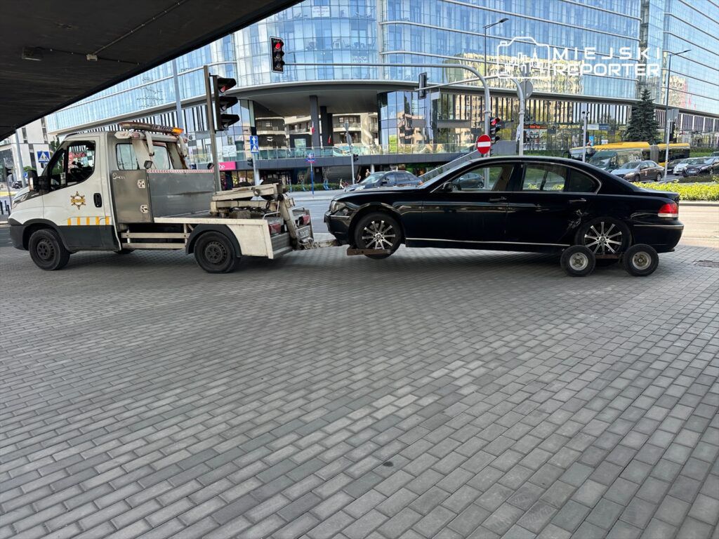 Laweta holująca czarny samochód osobowy na ruchliwej ulicy, obok nowoczesnych budynków i sygnalizacji świetlnej.