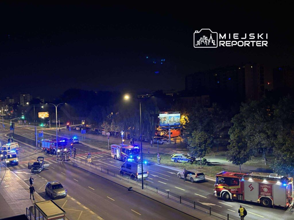Na ulicy zaparkowane pojazdy straży pożarnej i karetki, w tle policyjni funkcjonariusze prowadzący działania.