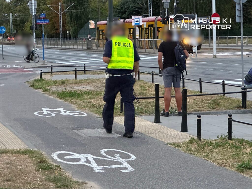 Policjant w kamizelce odblaskowej rozmawia z mężczyzną stojącym obok ścieżki rowerowej w miejskim otoczeniu.