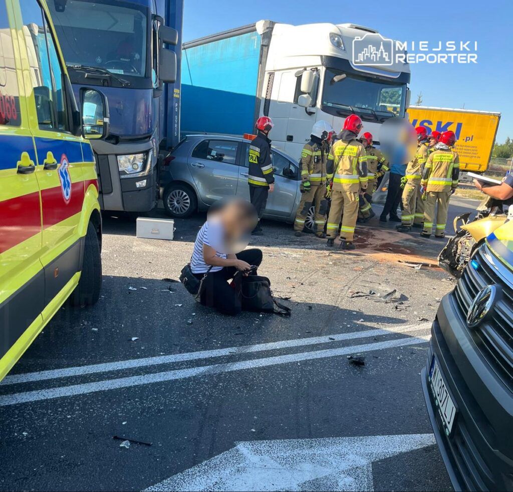 Kobieta w pasiastym T-shircie siedzi na jezdni obok wraku samochodu, podczas gdy strażacy udzielają pomocy poszkodowanym.