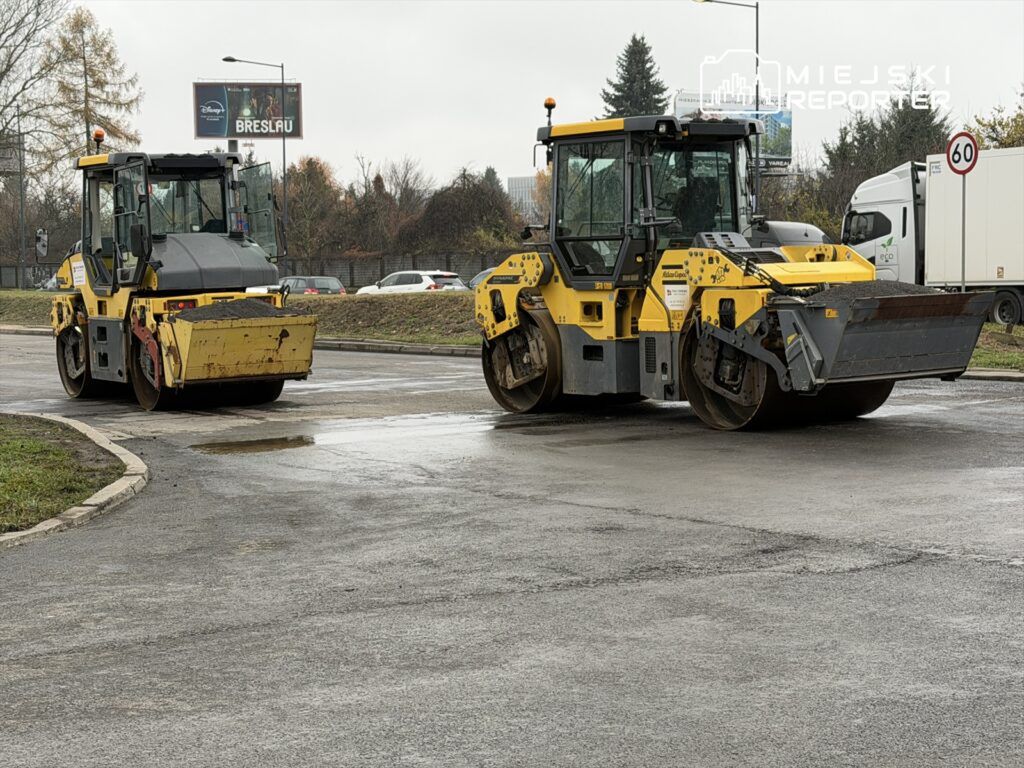 Dwa walce drogowe w żółto-czarnym kolorze stoją na mokrej nawierzchni, w tle widoczne samochody i billboard.