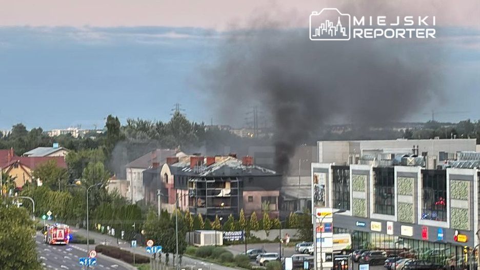 Czarny dym unoszący się z płonącego budynku mieszkalnego w tle, w pobliżu nowoczesnego centrum handlowego.