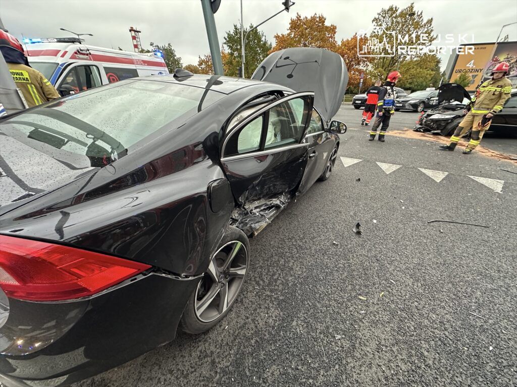 Czarny samochód osobowy z uszkodzeniami bocznymi stoi na skrzyżowaniu, obok pracują strażacy i ratownicy medyczni.