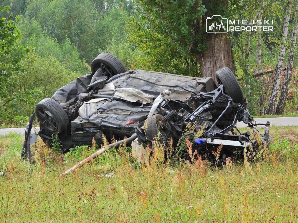 Przewrócony samochód leży na boku w zaroślach, otoczony wysoką trawą i drzewami.