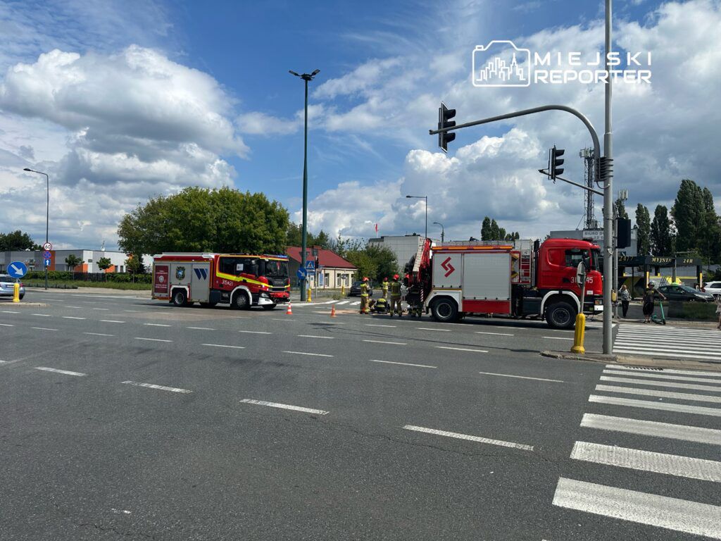 Na skrzyżowaniu ulicy znajduje się wóz strażacki oraz zespół strażaków, którzy prowadzą działania ratunkowe. Ulica jest częściowo zablokowana, a w okolicy widać oznakowanie i pojazdy czekające na wznowienie ruchu. W tle widoczne są budynki oraz drzewa.