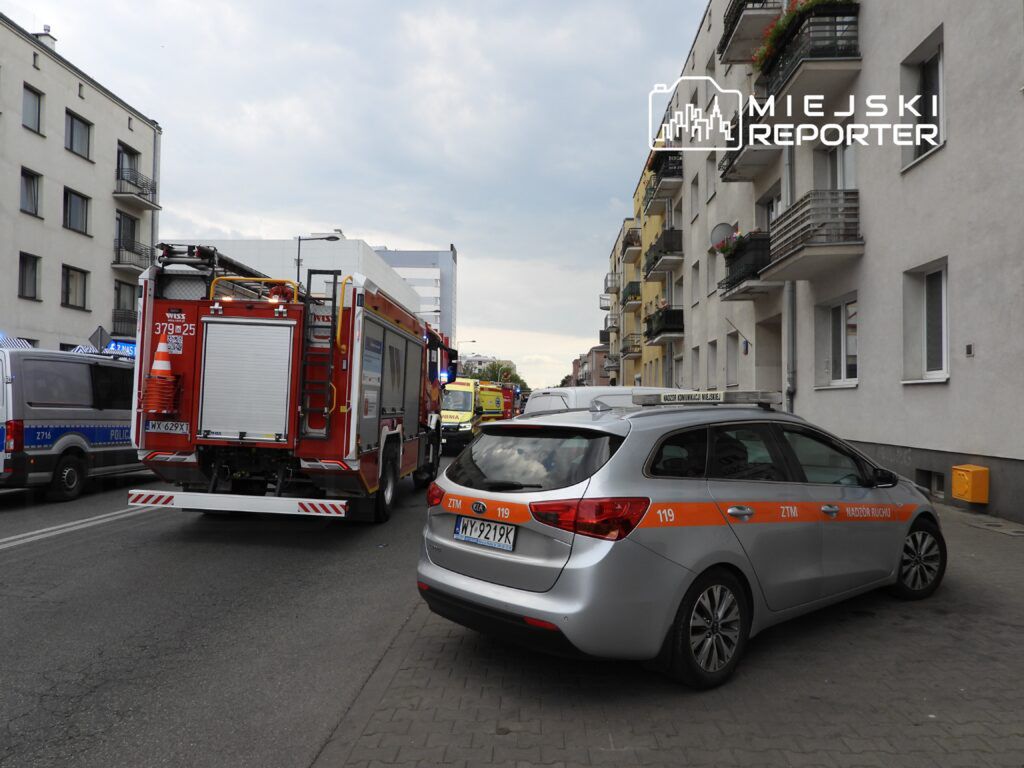 W tle widać strażacki wóz ratunkowy z włączonymi sygnałami świetlnymi, obok stoi radiowóz policyjny. Na ulicy zaparkowane samochody.