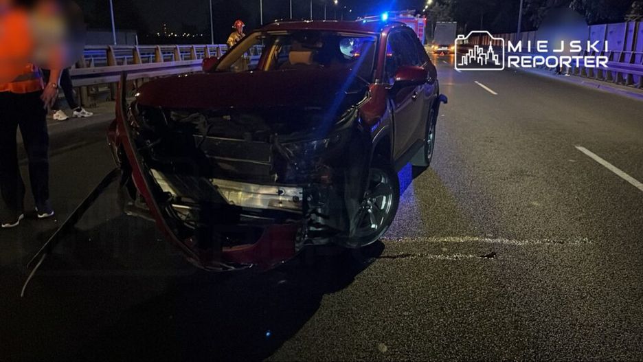 Uszkodzony samochód osobowy na drodze, obok stojący ratownicy w odzieży ochronnej przy włączonych sygnalizacjach świetlnych.