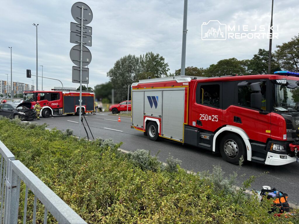 Dwa wozu strażackie na drodze, jeden z uszkodzonym samochodem, a obok ekipa ratunkowa zabezpiecza miejsce zdarzenia.